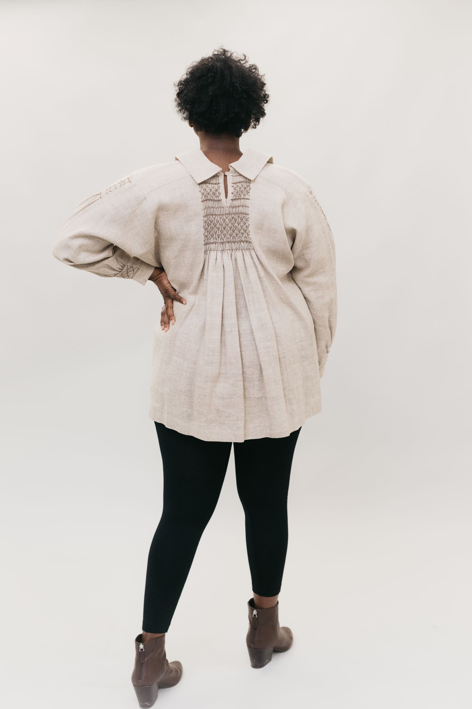 Back of African American woman standing in front of a white studio backdrop with hand on her hip wearing 221 English Smock Frock, smocked in the back, with black leggings and brown heeled Chelsea boots.