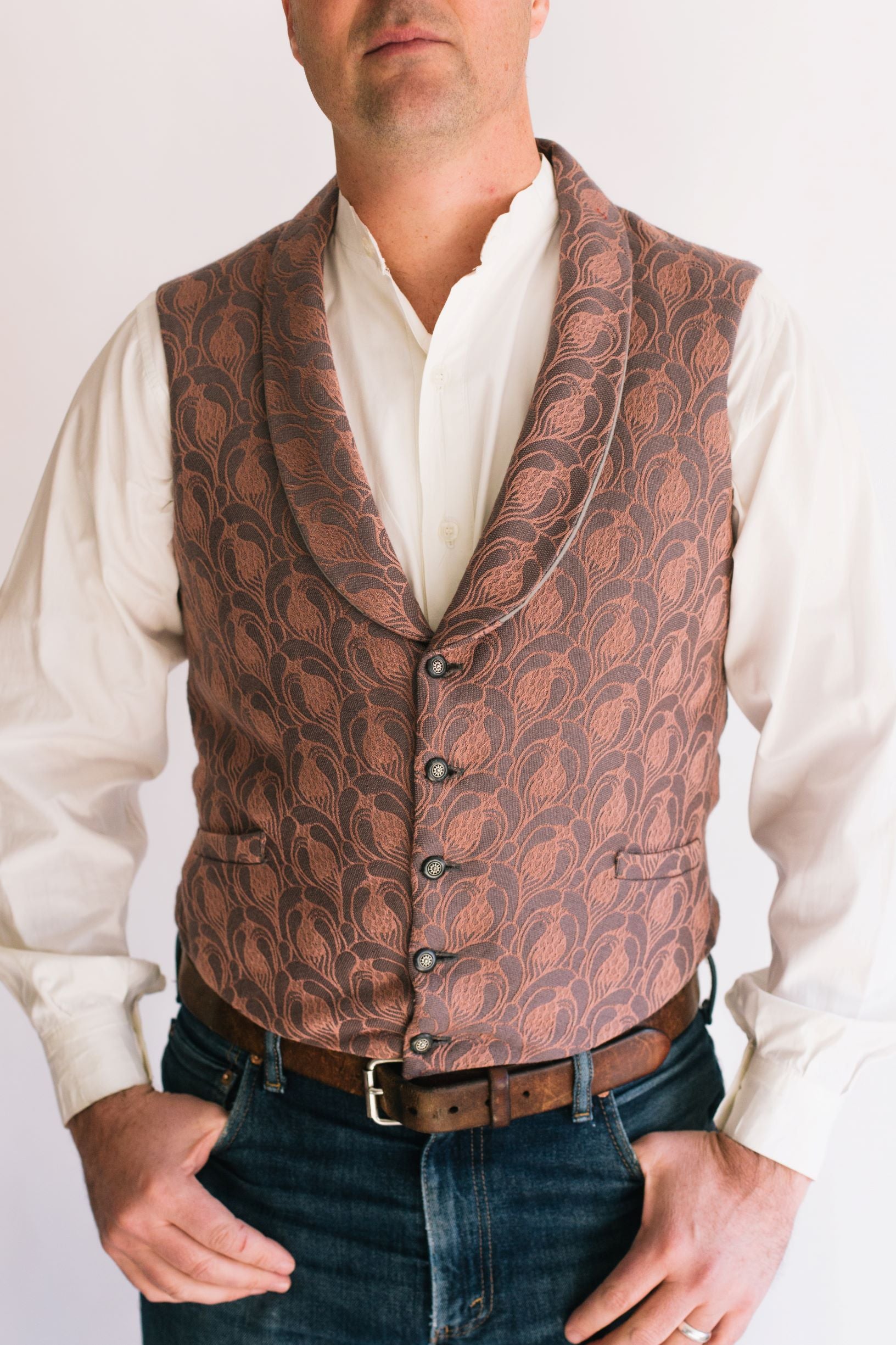 Man standing in front of a white studio backdrop, wearing 222 Vintage Vests with shawl collar. long sleeve shirt worn underneath with thumbs in pockets