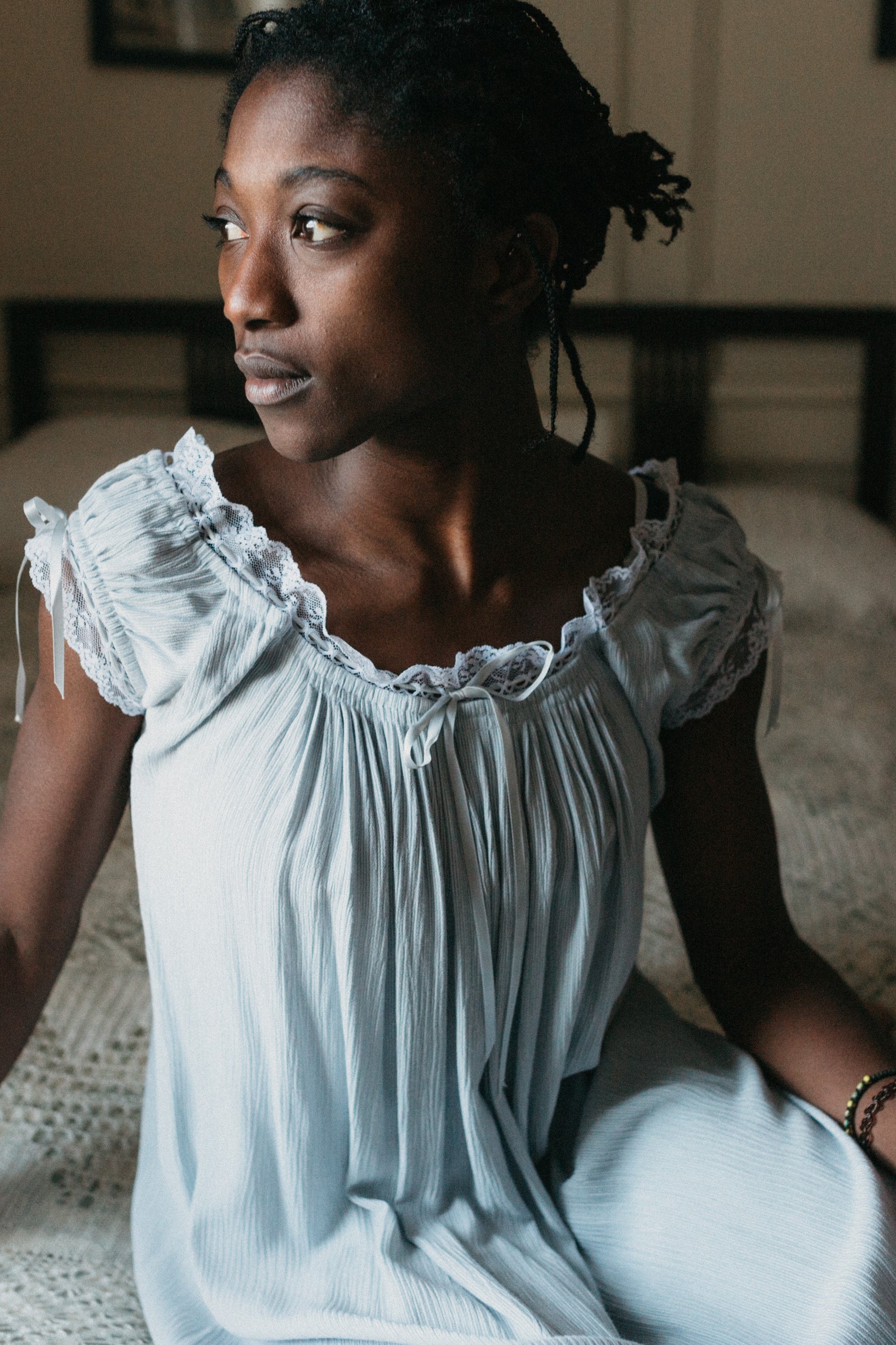 African woman sitting on a bed looking to the left wearing 223 A Lady's Chemise, View B with neckline sitting on the shoulders, self fabric ruffle and casing for the ribbon drawstring.