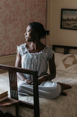 African woman sitting at the foot of a bed with both legs folded underneath her, looking to the left wearing 223 A Lady's Chemise.