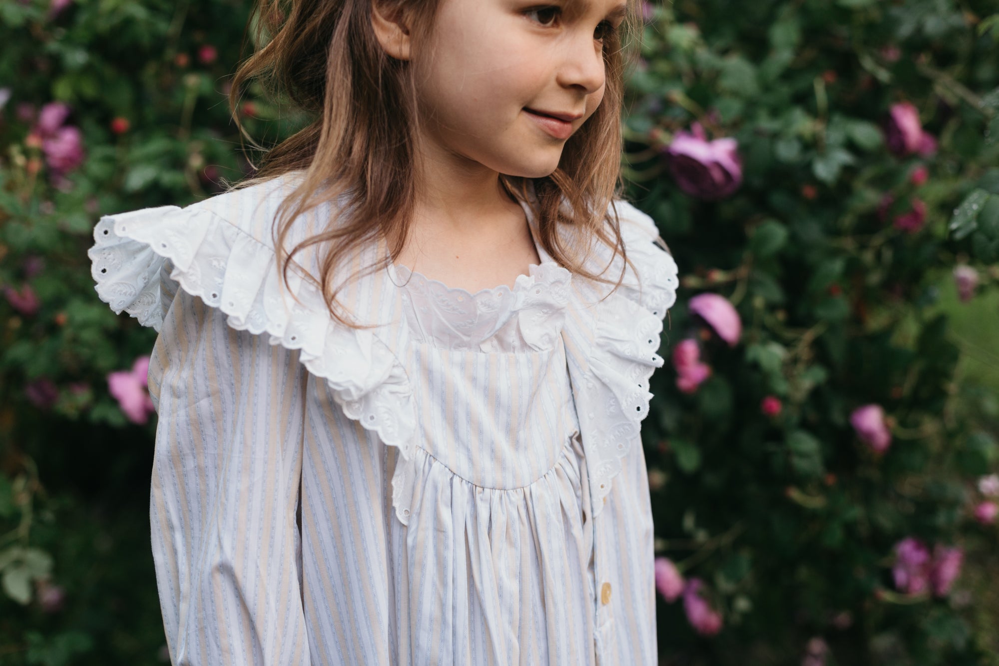 Brunette little girl surrounded by greenery and purple roses close up of her wearing 225 Childhood Dreams.