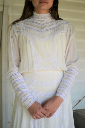 Young brunette woman standing in front of a white wall wearing 227 Edwardian Bridal Gown and Dress, with lacy detail, on the bodice, skirt, and cuffs.