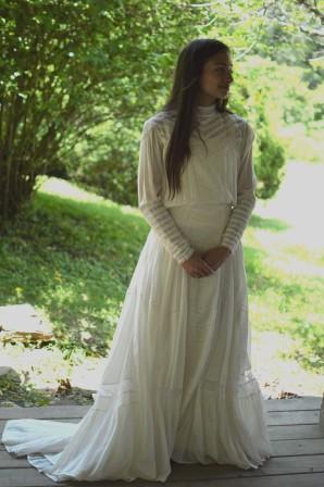Young brunette woman standing on a wooden platform surrounded by greenery, wearing 227 Edwardian Bridal Gown and Dress. 