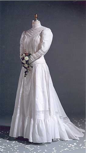 Mannequin wearing 227 Edwardian Bridal Gown and Dress standing in front of a gray studio backdrop, holding a bouquet, with white flower petals sprinkled on the floor.