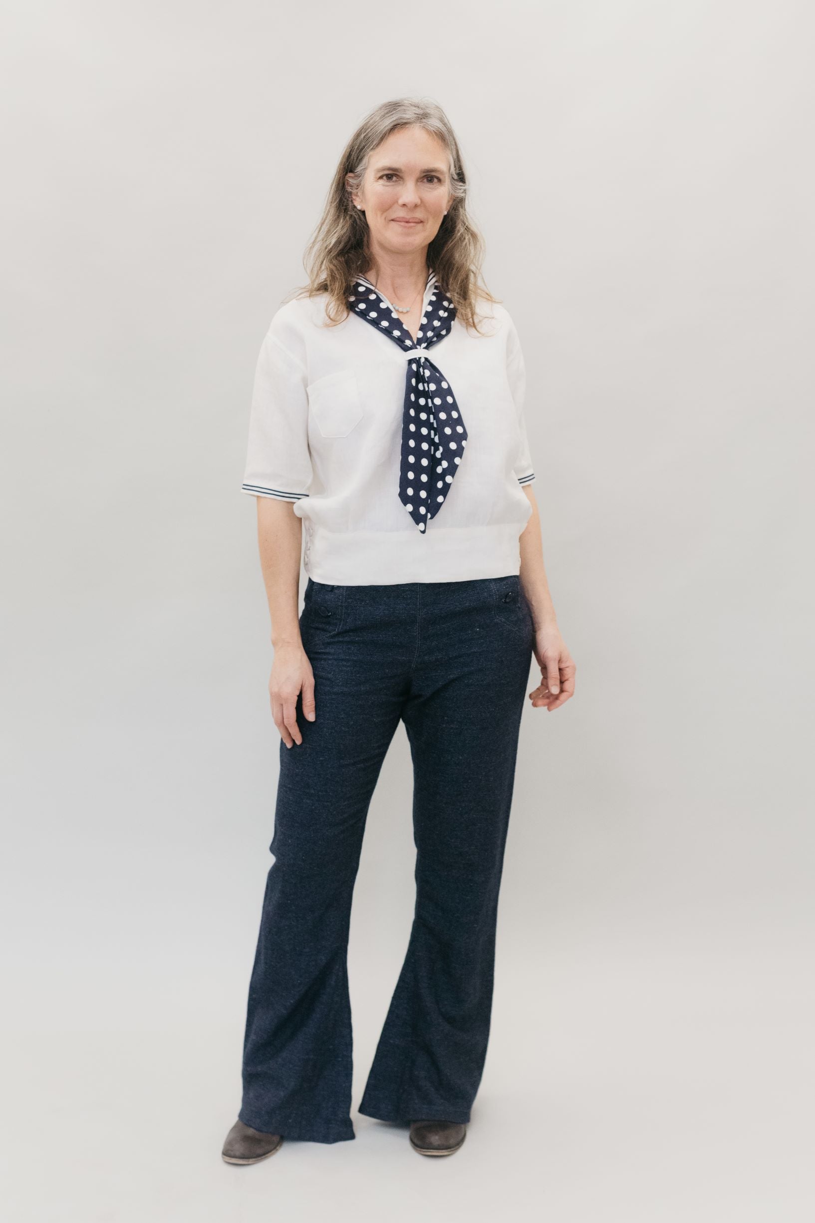 Woman smiling and standing in front of a white studio backdrop wearing 229 Sailor pants and 211 Two middies with scarf. 