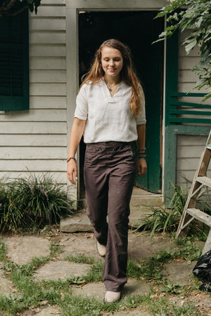 Young girl walking in front of a doorway, looking down smiling wearing a white top, beige shoes and 229 sailor pants.