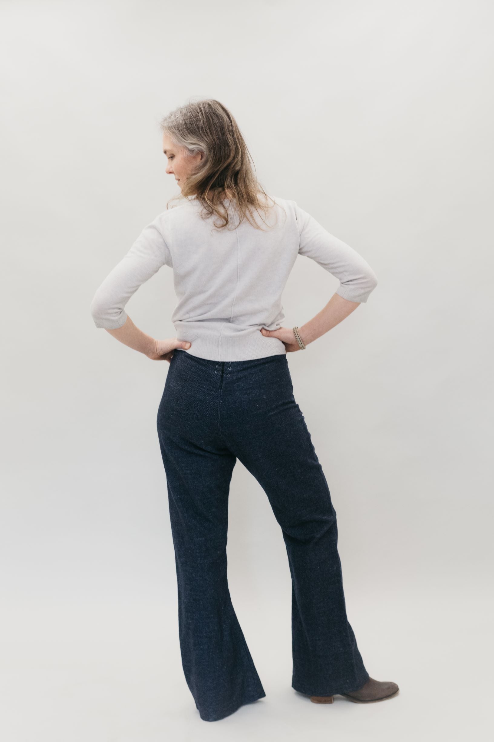 Back view of woman standing with both hands on hips, head turned to the side, wearing 229 Sailor pants and a white long sleeve.