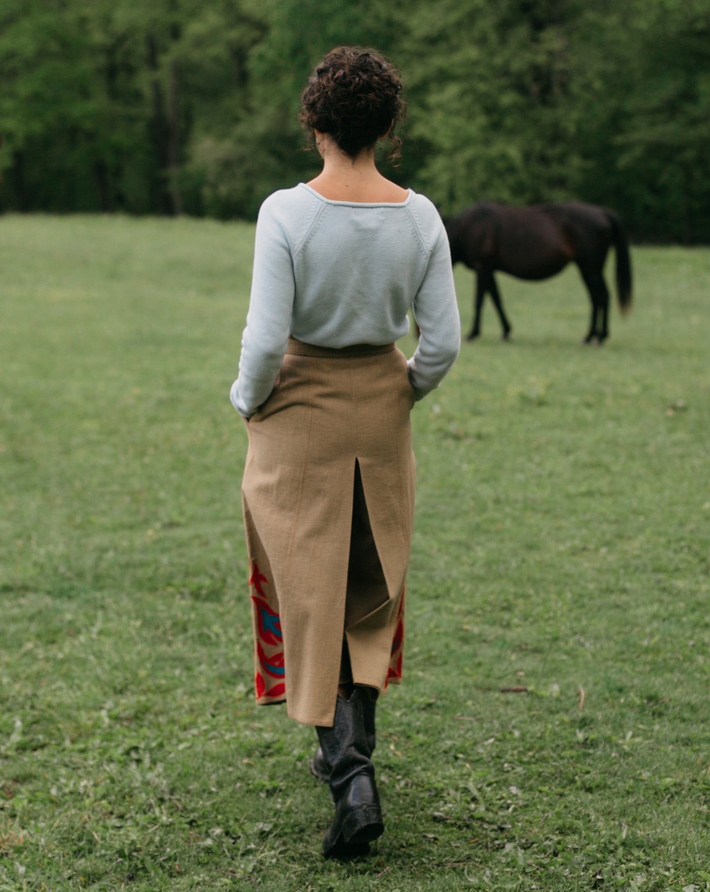 Back view of woman walking surrounded bu greenery with hands in the pocket of the culotte 231 Big Sky Riding Skirt, with a horse in the background.