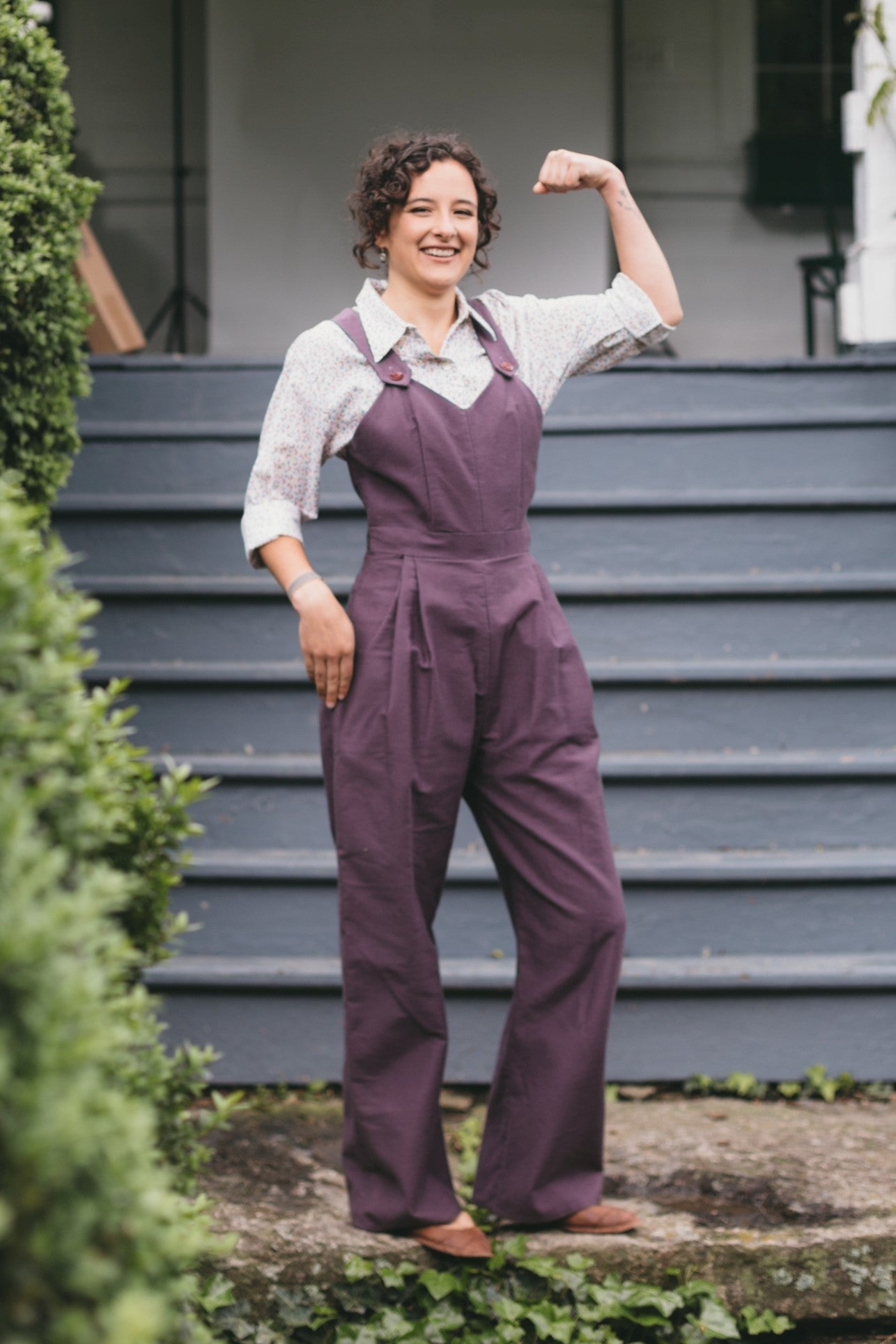 Young brunette white woman standing, smiling white showing off her arm muscles, wearing the 240 Rosie the Riveter overalls. 