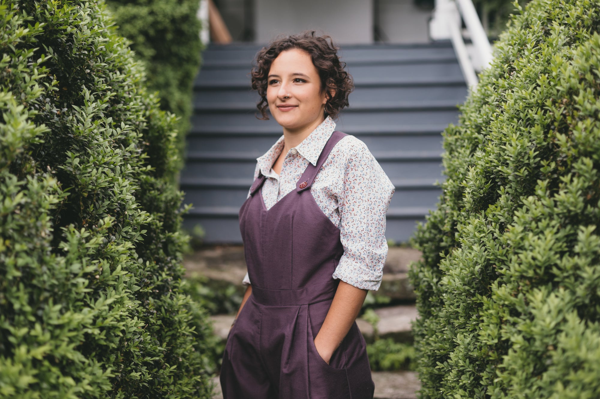 Young brunette woman turned slightly to the left wearing the Rosie the Riveter overalls with hands in the pockets surrounded by greenery. 