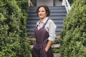 Young brunette woman turned slightly to the left wearing the Rosie the Riveter overalls with hands in the pockets surrounded by greenery. 
