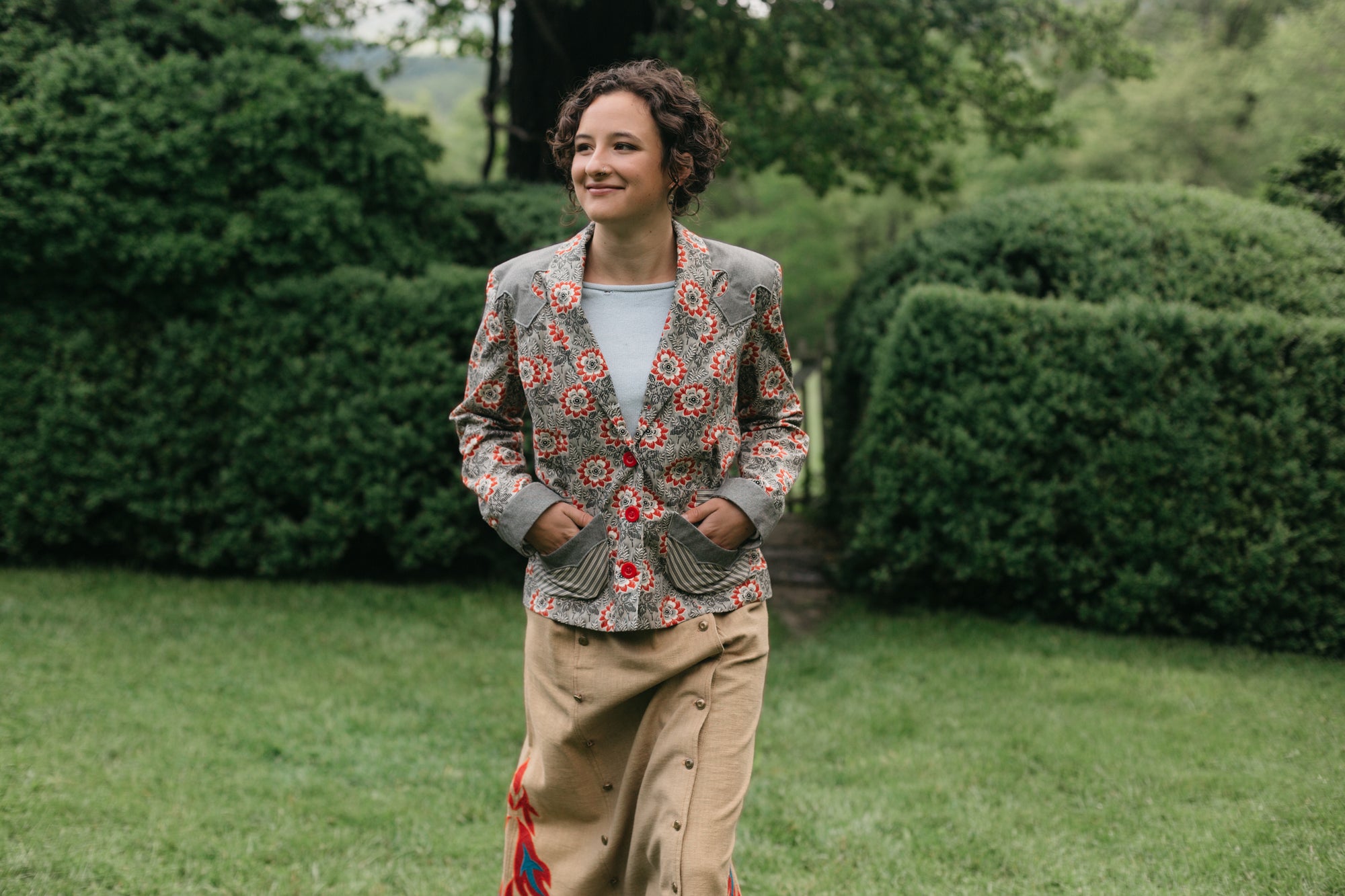 Young brunette woman smiling walking surrounded by greenery wearing the 242 Rodeo Cowgirl Jacket with hands in the pockets. 