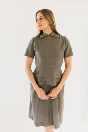 Woman standing with hands in front of her looking to the side in a tan colored version of #249 1930s Day Dress.  She is wearing view A. View A features round neck with collar points and peplum cleverly cut on the straight grain but with the slimming look of a bias cut.