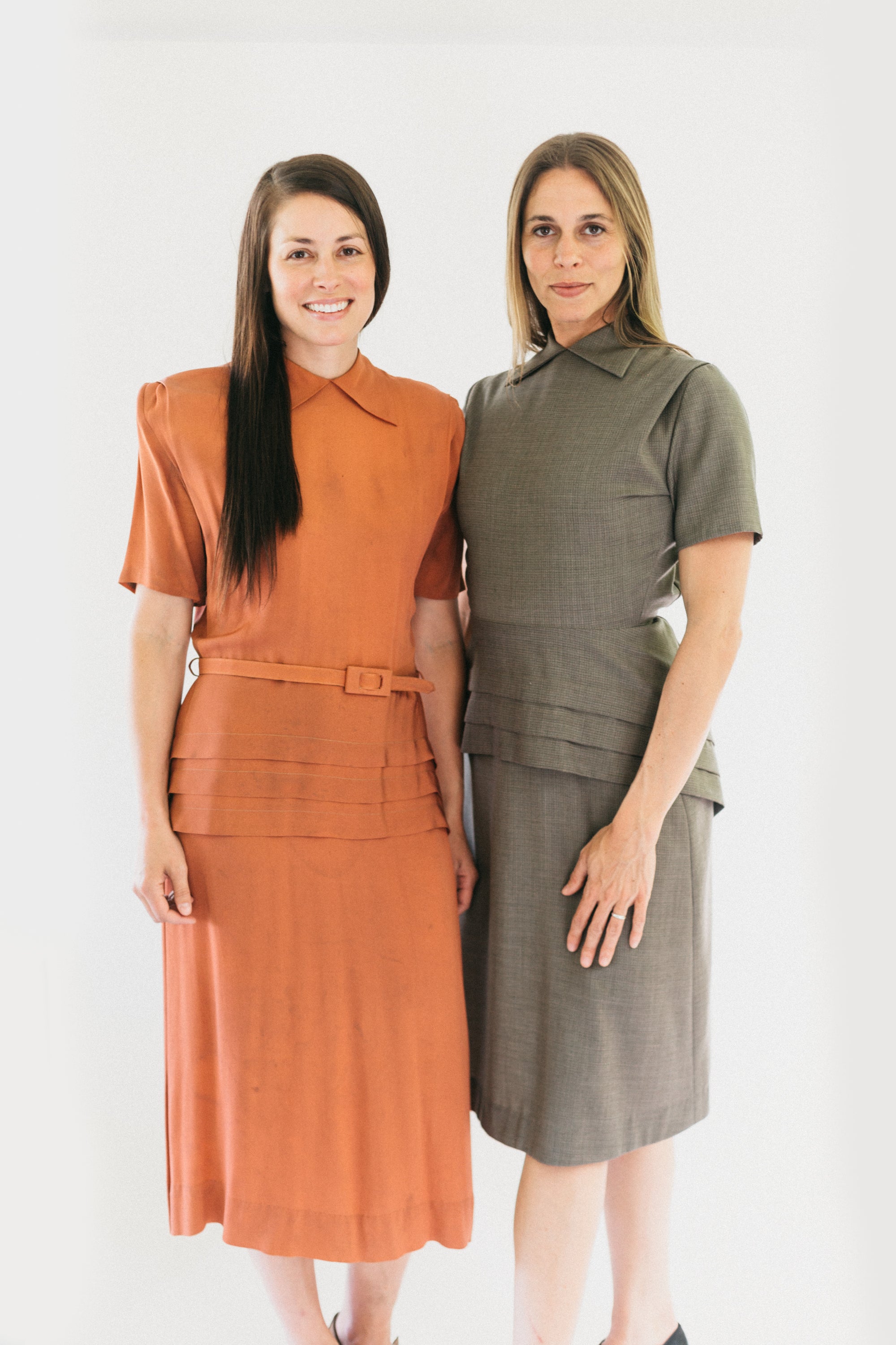 Two women standing in 1930s Day Dresses.  Both are looking forward and standing in front of a white backdrop.  Dress on left is a light salmon color.  On right is a tan color.  Botha re wearing view A.  View A features round neck with collar points and peplum cleverly cut on the straight grain but with the slimming look of a bias cut.