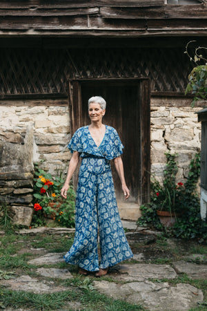 Woman walking in beach pyjamas outdoors in front of stone building with flowers by door.