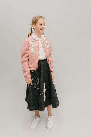 A young blonde girl with pig tails wearing #256 At The Hop smiling and standing in front of a white studio backdrop.