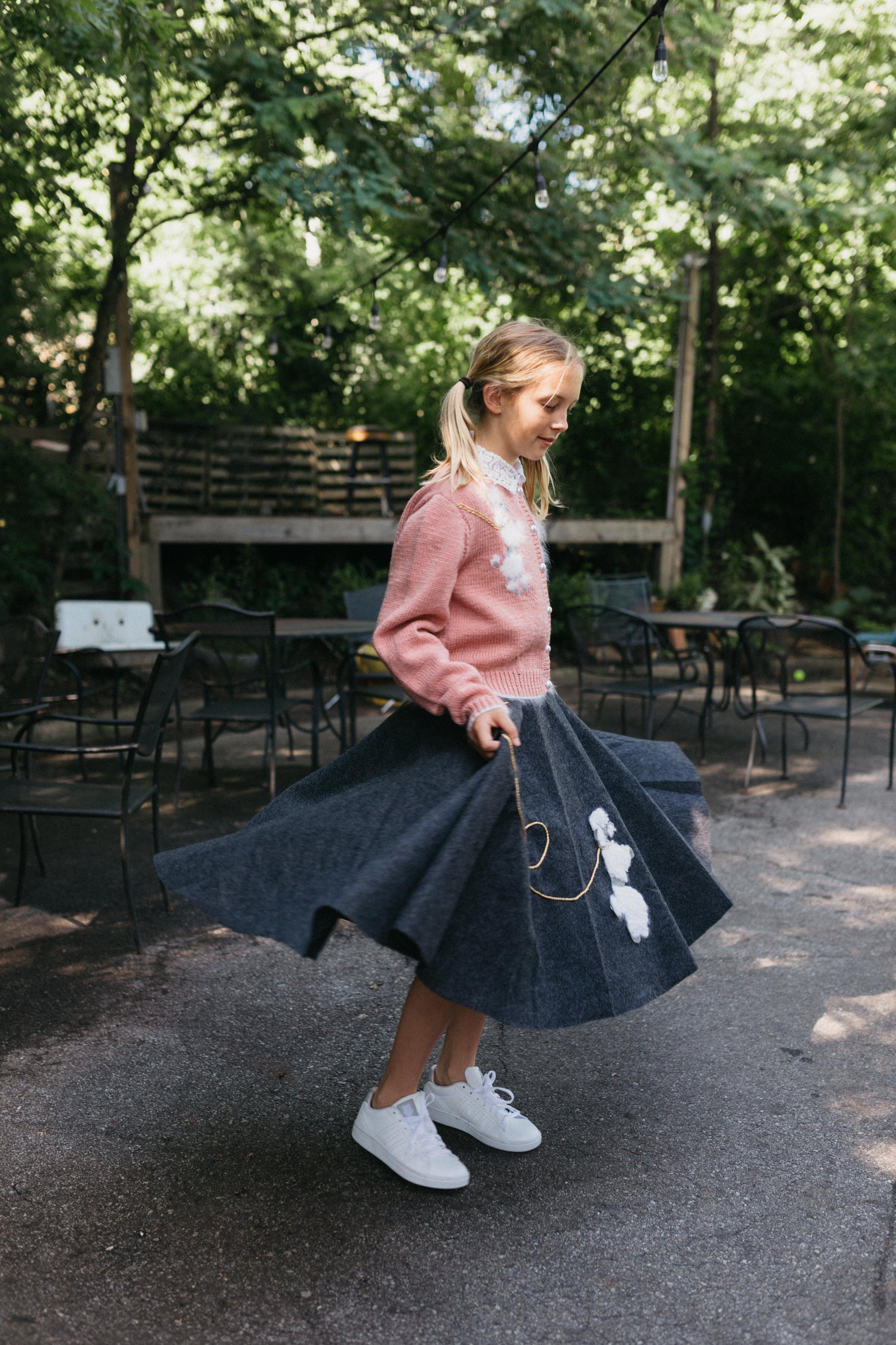 Young blonde girl twirling surrounded by greenery and chairs behind her, wearing At The Hop skirt and Sweater.