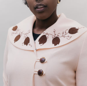 Black woman wearing a pink wool coat with brown leaf applique and embroidery on cuffs and collar.