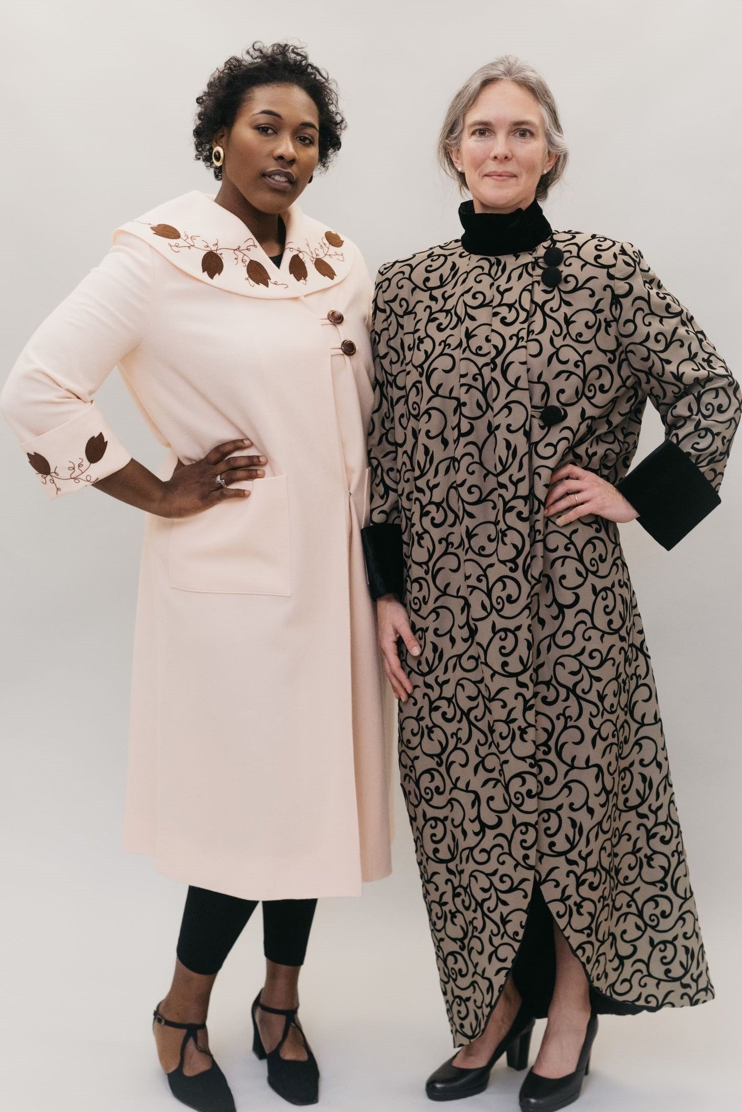 Two women wearing Spectator Coats. Coat on right is brown and black with black cuffs and high collar. Woman on left is wearing a pink wool coat with brown applique at cuffs and collar.