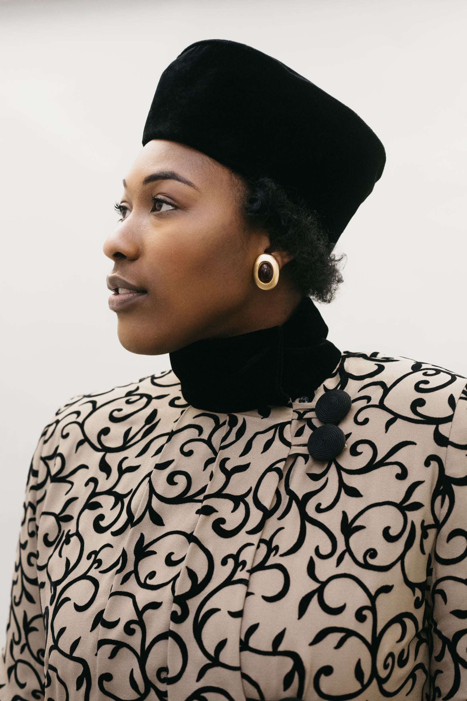 Close up of a Black woman wearing a brown and black coat with black velvet collar and turban hat.