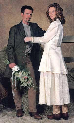 Man standing with flowers in his right hand with woman on his left  wearing a white dress and coat looking at the viewer about to button his Countryside Frock Coat.