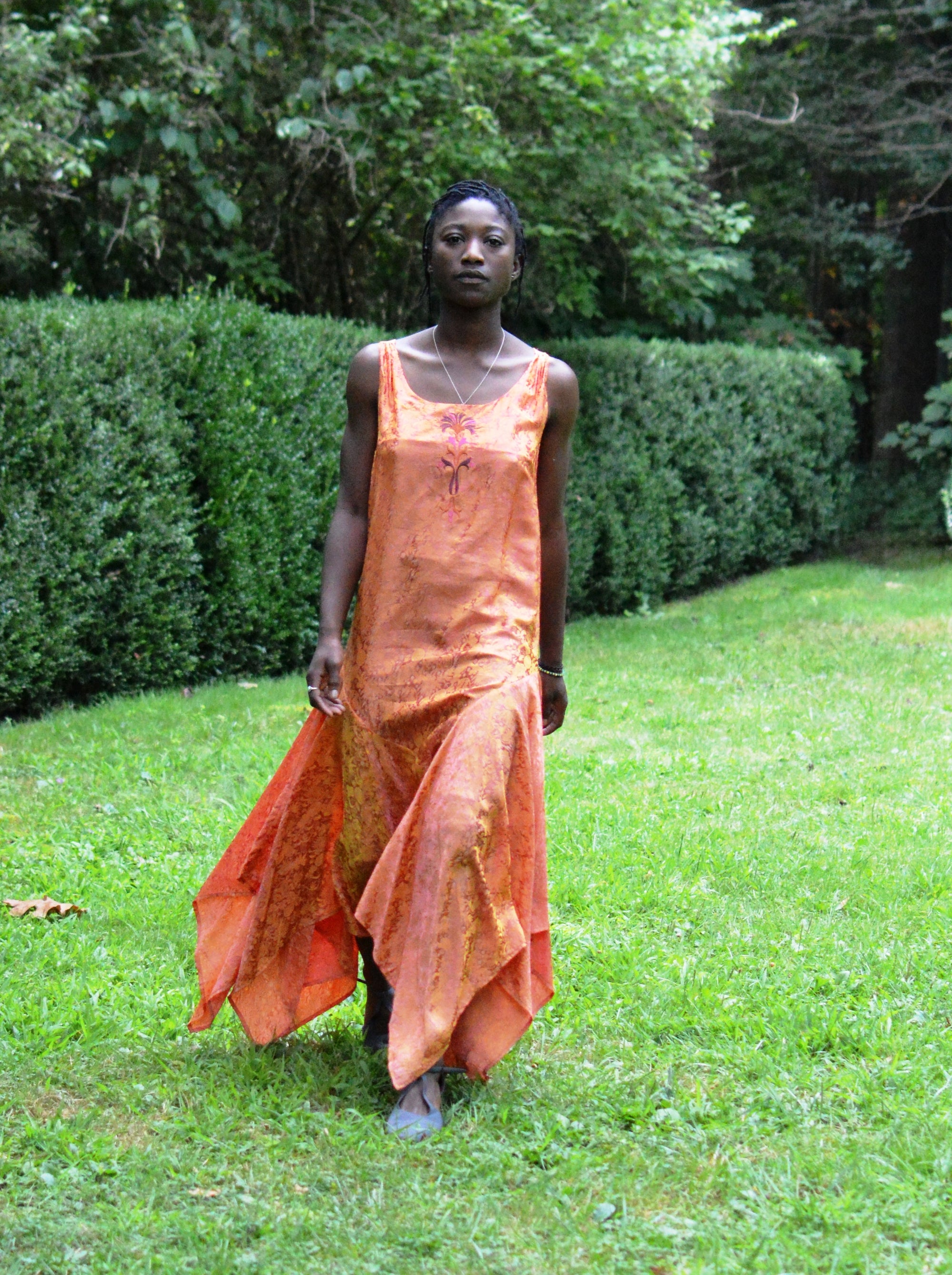 Young African woman standing surrounded by greenery wearing #264 Monte Carlo Dress in orange. 