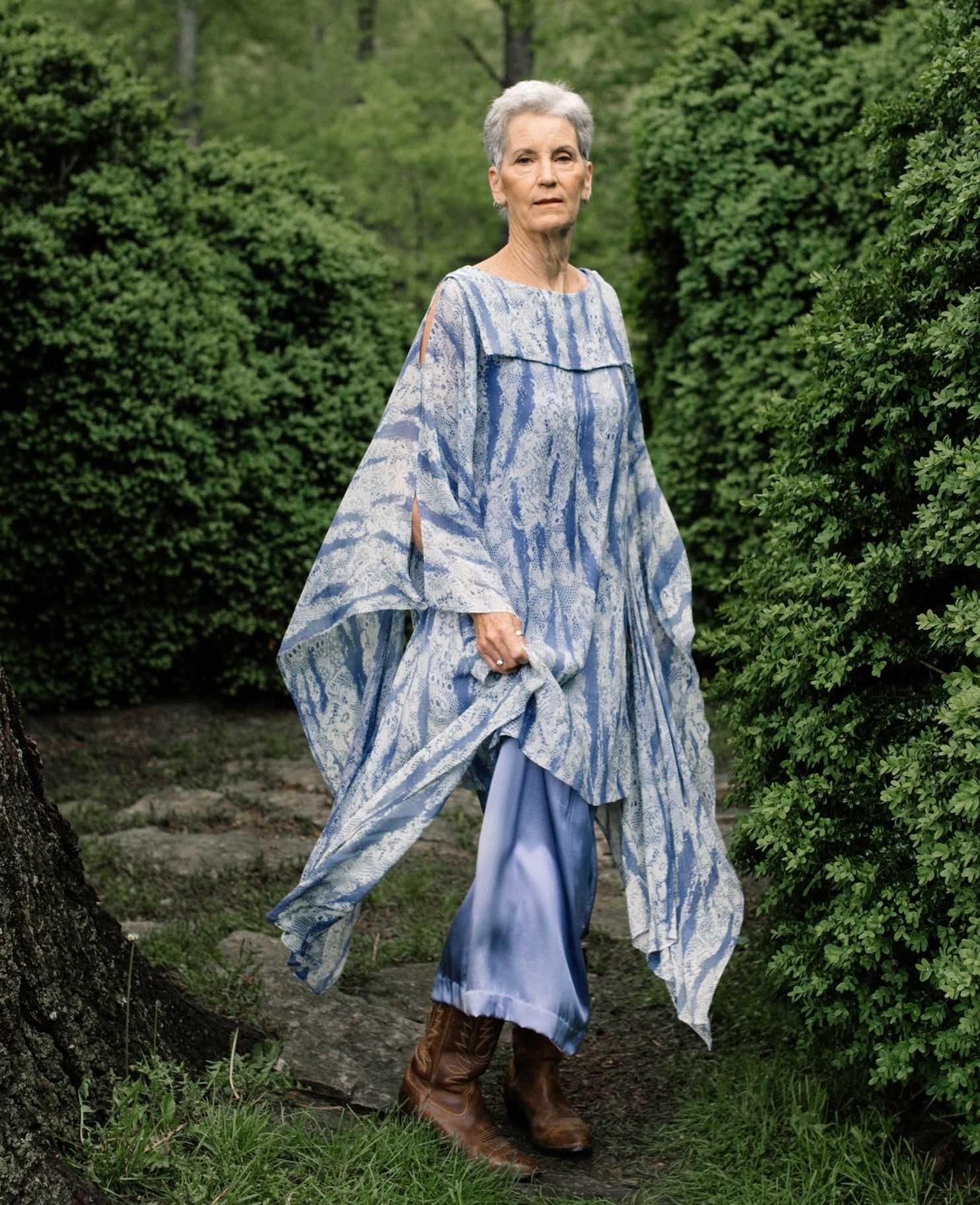 Older white woman with short grey hair standing surrounded by greenery,wearing brown boots and the Greek Island Dress Tunic displaying the flare of the free- floating sleeve panels. 