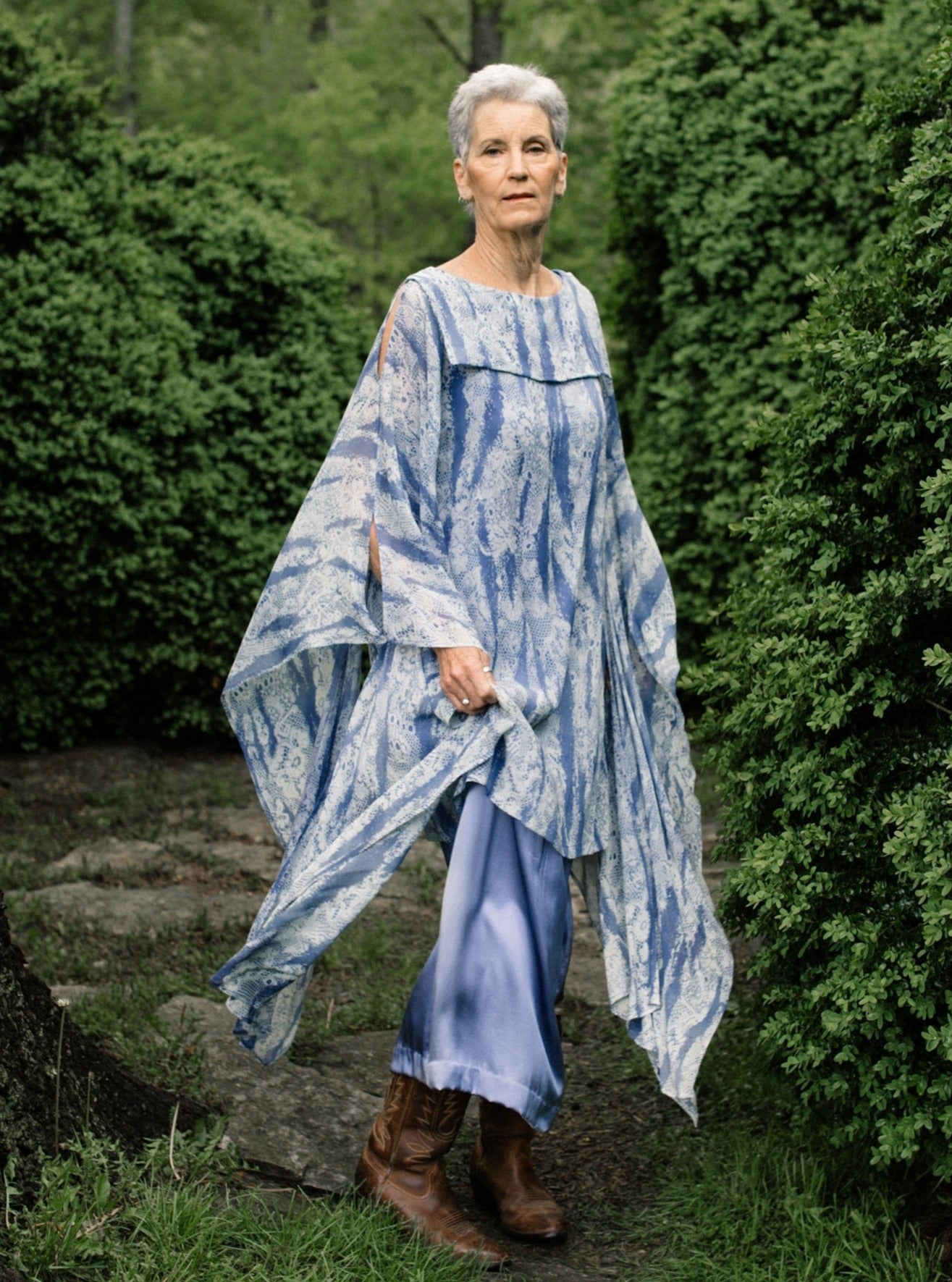 Older white woman with short grey hair standing surrounded by greenery,wearing brown boots and the Greek Island Dress Tunic displaying the flare of the free- floating sleeve panels. 