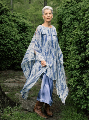 Older white woman with short grey hair standing surrounded by greenery,wearing brown boots and the Greek Island Dress Tunic displaying the flare of the free- floating sleeve panels. 