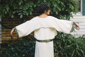 Young Brunette White woman wearing the Greek Island Dress with Jacket, showing the back view of the Dress with jacket, extending both arms showing the center back godet that adds flare.