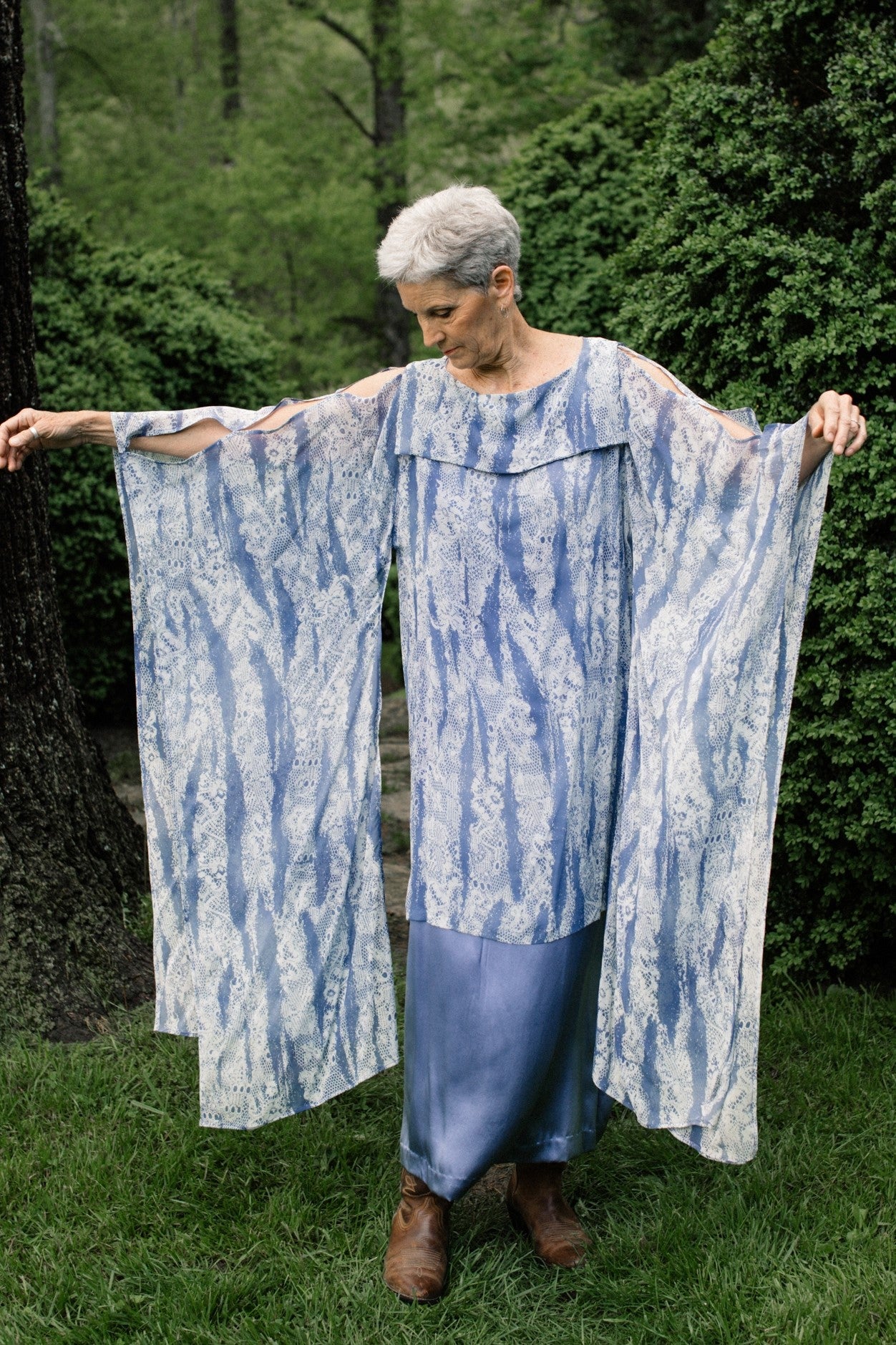 Older White woman with grey short hair standing surrounded by greenery looking down, wearing the #266 Greek Island Dress Tunic, extending both arms out showing the free-floating sleeve panels.