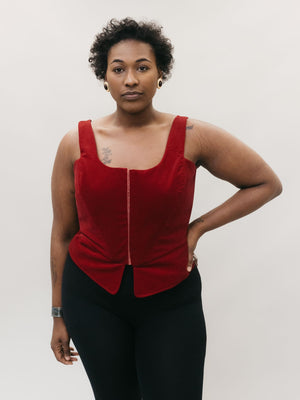African American woman wearing red #267 M' Lady's Corset with a scoop neck and black pants, standing in front of a studio white backdrop with her left hand on her hip