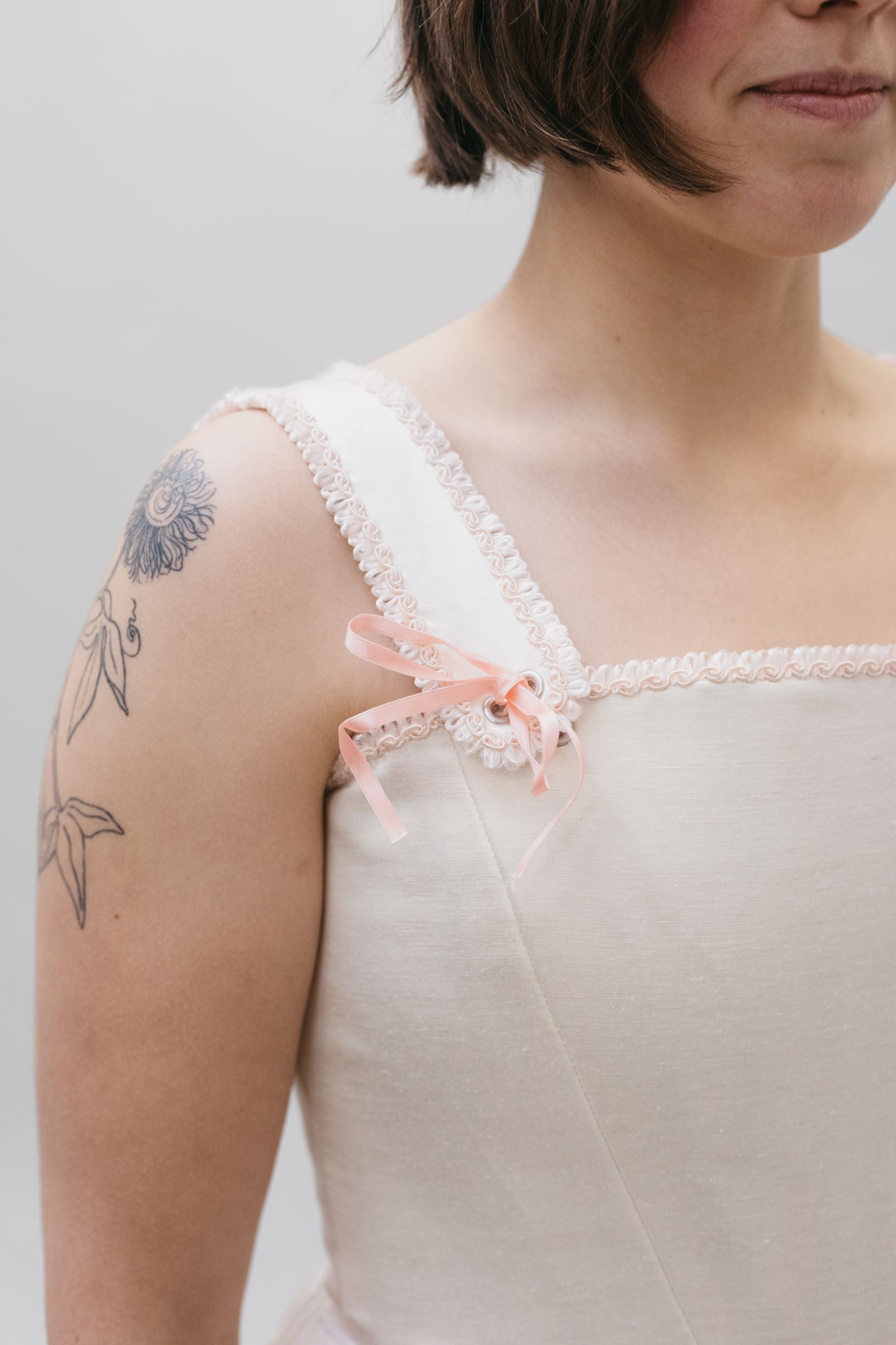 Close up of the square neck Lady's Corset strap withe a pink bow ribbon at the base of the strap on white woman with dark hair and tattoo.  Ribbon tie detail for Corset top.