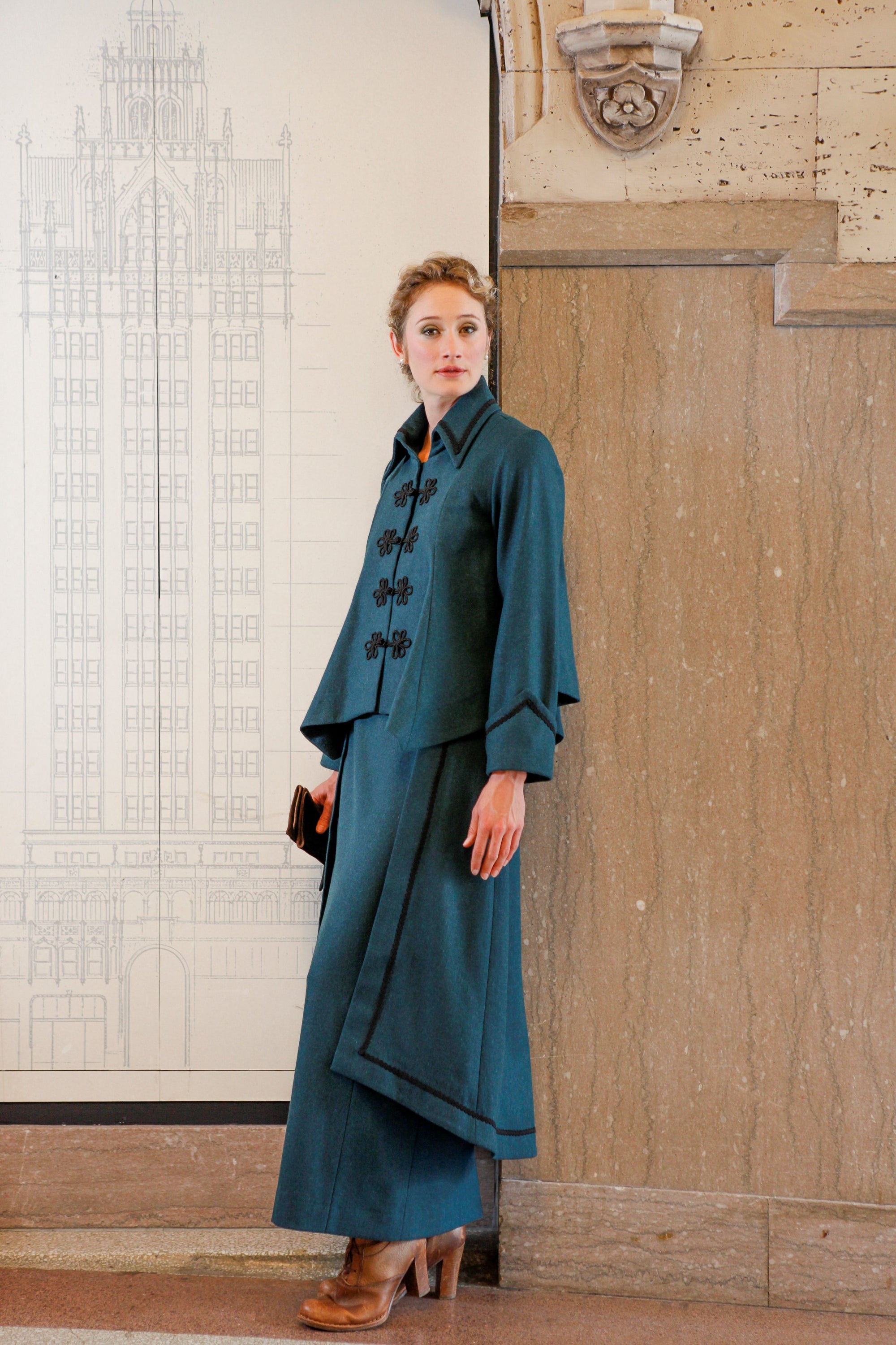 Young blonde white woman standing in between a black and white architectural drawing and a brown marble wall, wearing #268 Metropolitan Suit in blue and wearing light brown heels holding a walled in her right hand.