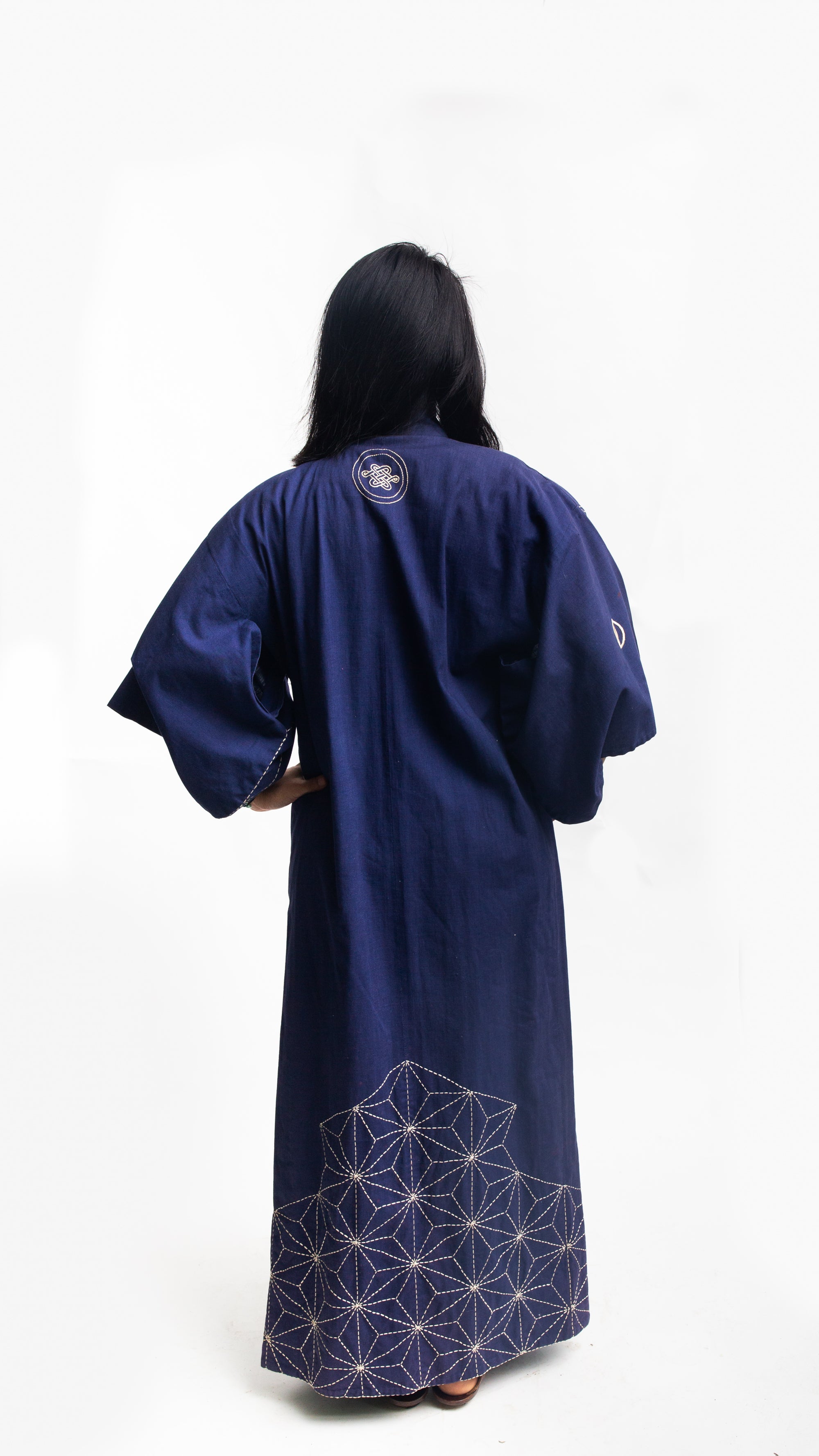 Asian American woman wearing a navy embroidered kimono standing in front of a white background. Back view with sashiko embroidery.