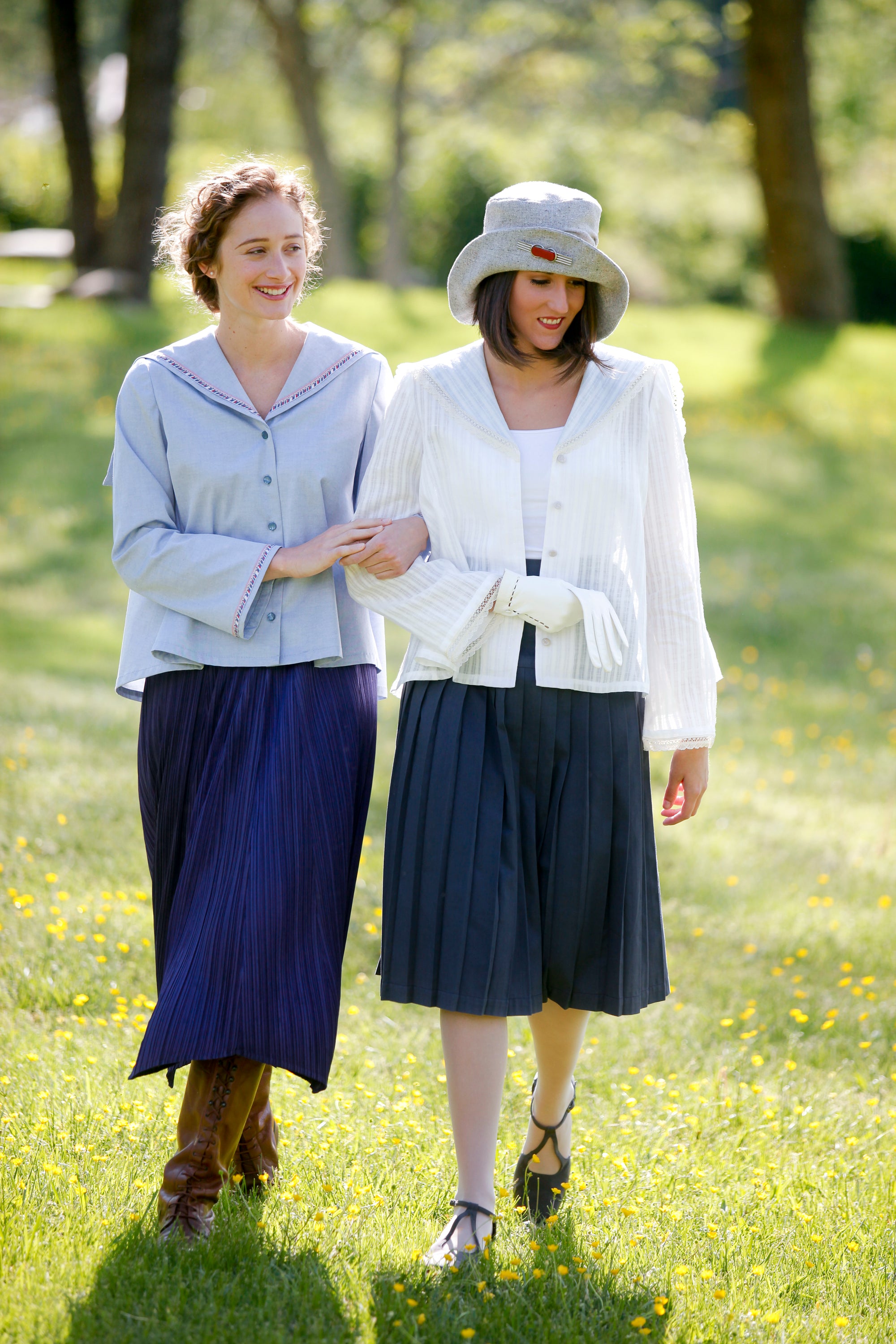 Two  women walking linking arms, surrounded by greenery. Woman on the left wearing #270 Metro Middy Blouse in blue and long skirt. Woman on the right is wearing a hat with the Metro Middy Blouse in white and a knee length skirt. 