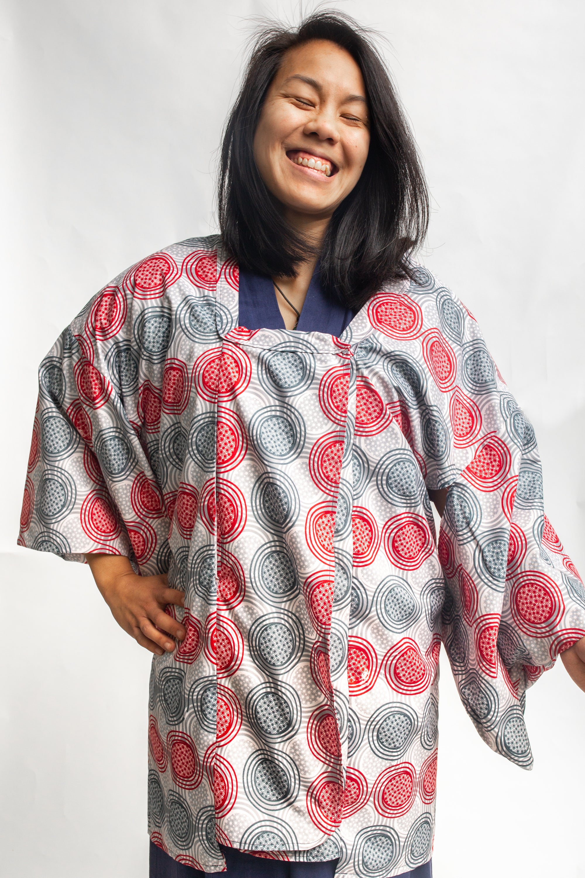 Smiling woman wearing a red and grey spiral printed ankara fabric made into a Michiyuki over a blue cotton kimono.  In front of a white back drop. 
