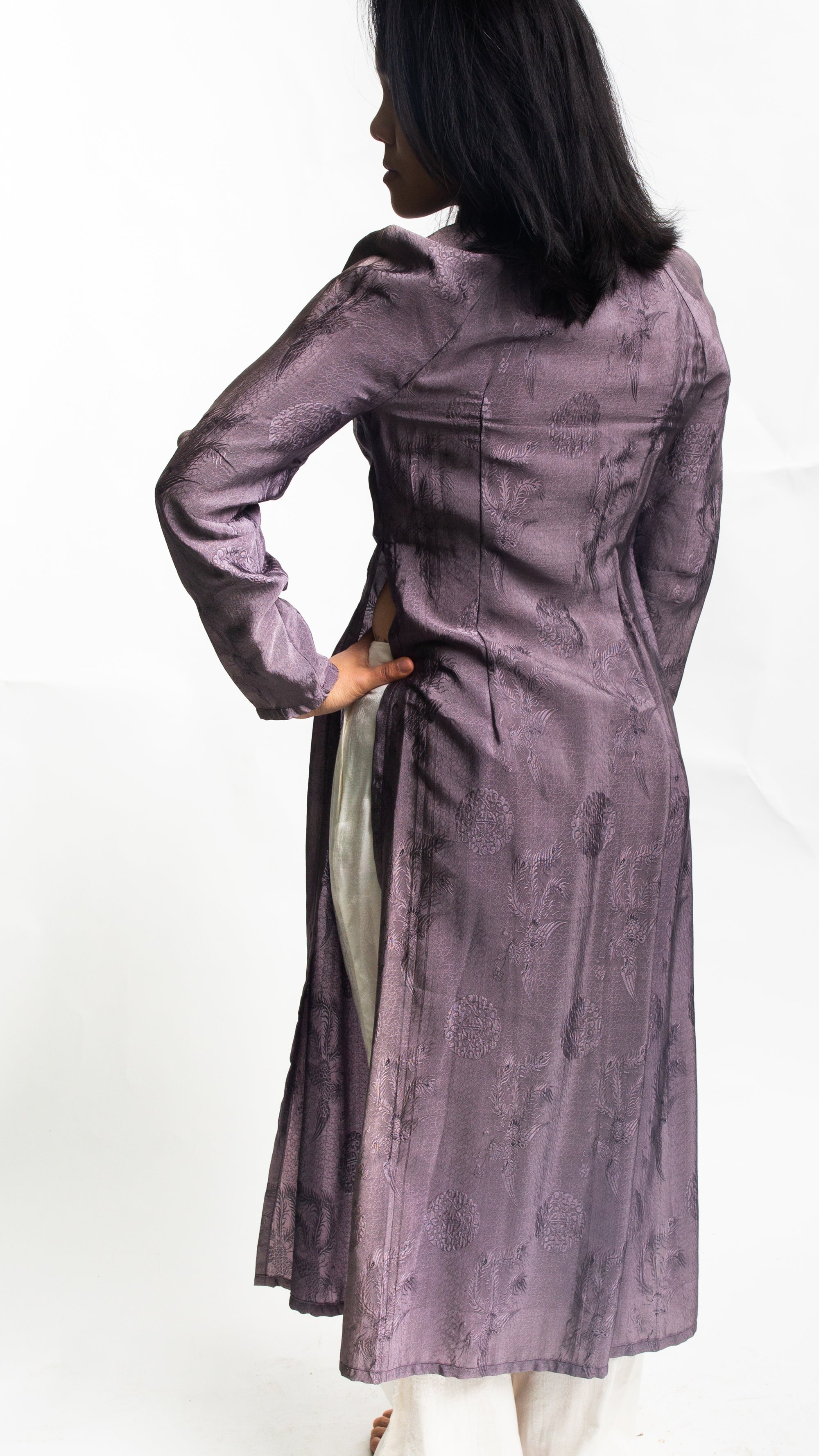 Back View of Vietnamese American woman wearing a purple silk Ao Dai tunic with white pants in front of white backdrop.