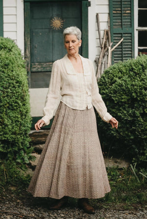 Older woman standing in front of a door with arms our surrounded by greenery wearing 210 Armistice Blouse and 209 Walking Skirt.