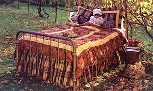 A girl sitting in bed underneath the covers outside surrounded by greenery with the 302 Victoria's Boudoir set, quilt cover, pillow shams, dust ruffle, throw pillows. 