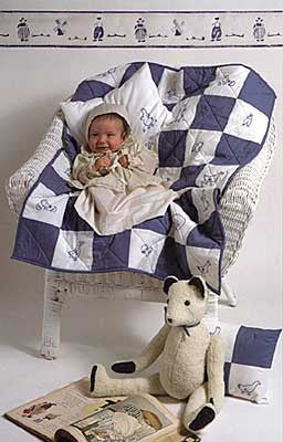 Black and white pen and ink drawing by Gretchen Schields. A baby sitting on a bed wearing 304 Nursery Days Bed Jacket and bonnet, and sitting on the quilt next to the teddy bear and pillow sham. 