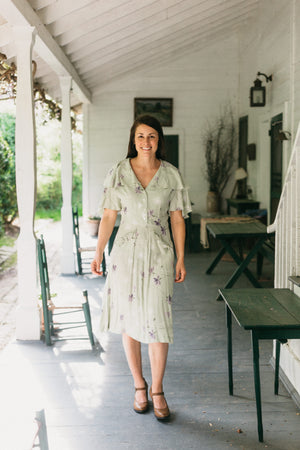 Woman walking towards the camera out doors on a sun lit porch.  There are tables in the foreground and chairs and a table in the background.  She is wearing a mint green and lavender floral dress.  Dress is View B. View B is more casual, with buttoned front, flounce collar and sleeves, and eyelash pockets.