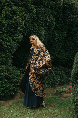 Young white woman with blonde hair walking face down in front of green bushes, wearing a dark blue dress under the Poiret Cocoon Coat