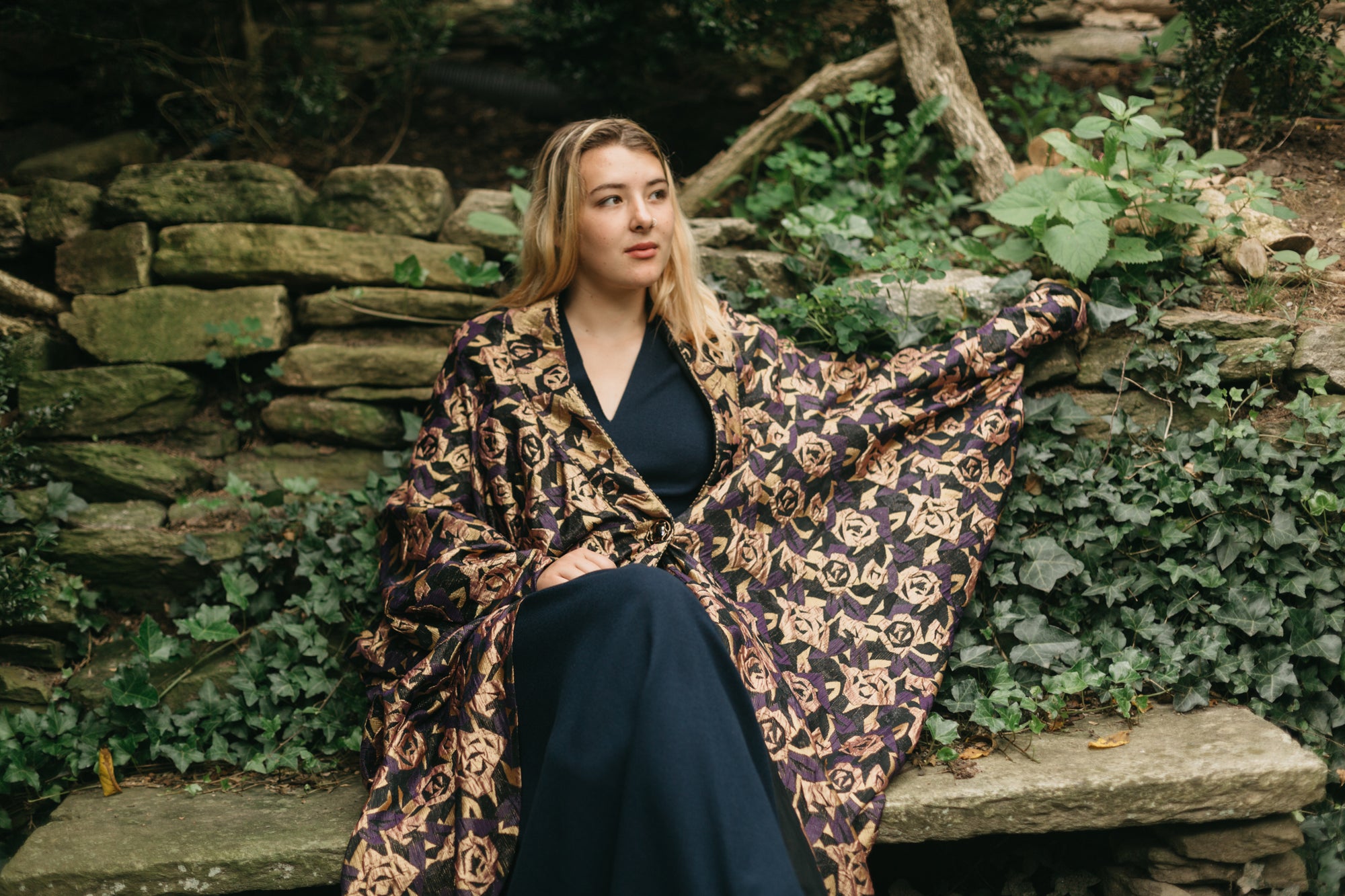 Young white blonde woman sitting on a stone bench with left hand stretched on a stone wall wearing the Poiret Cocoon Coat