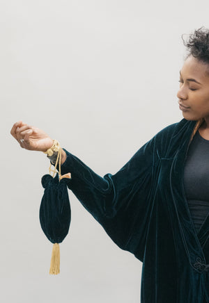 An African America woman wearing a velvet dark green Poiret Cocoon Coat against a studio white backdrop, holding a dark green velvet drawstring bag with gold cording.