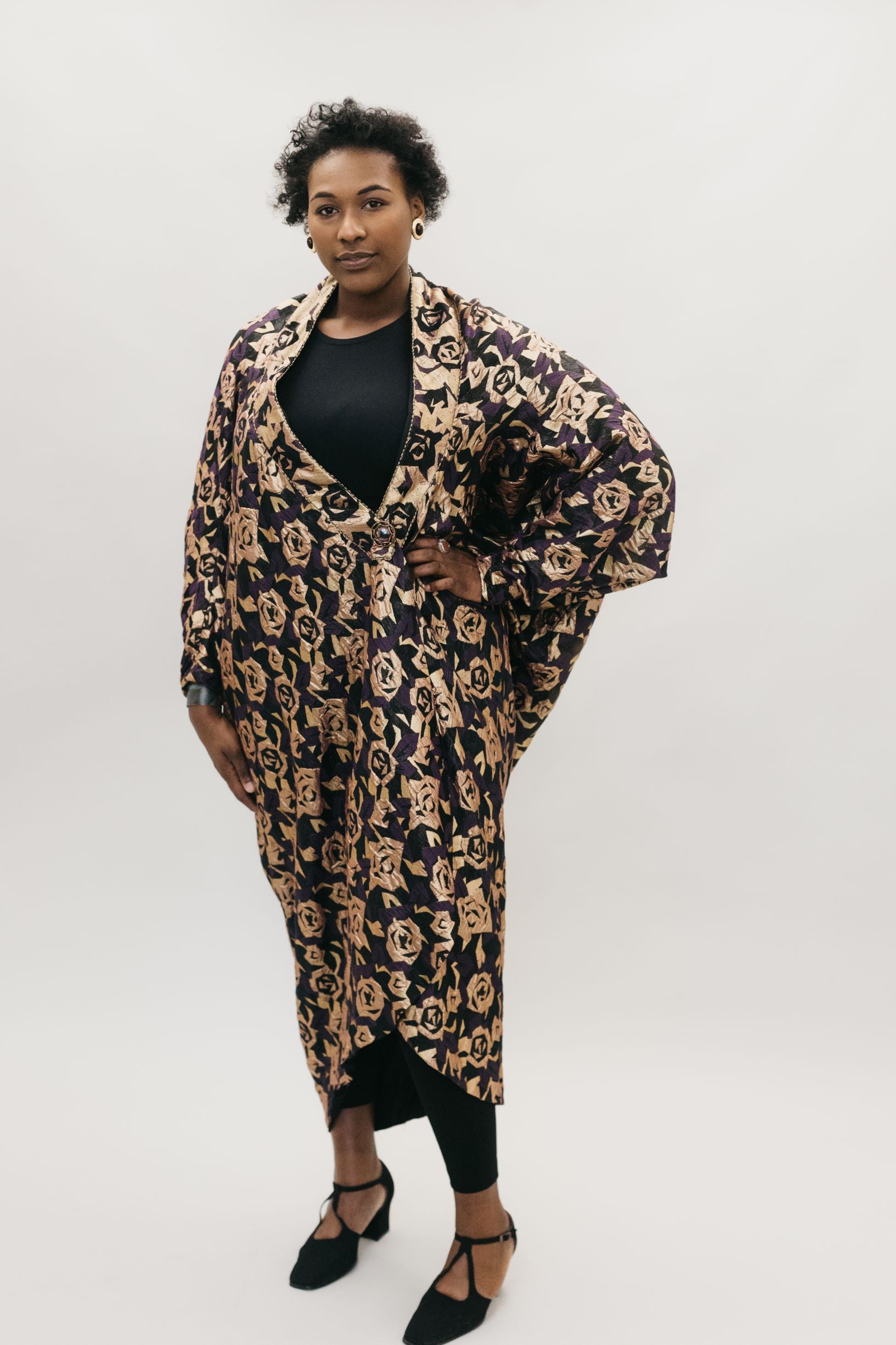 African American woman standing with left hand on her hipin front of a studio white backdrop wearing the 503 Poiret Cocoon Coat in a silk charmeuse fabric with gold roses as the pattern. She is also wearing black closed toed shoes. 