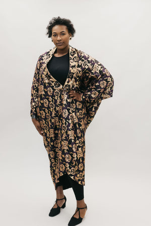 African American woman standing with left hand on her hipin front of a studio white backdrop wearing the 503 Poiret Cocoon Coat in a silk charmeuse fabric with gold roses as the pattern. She is also wearing black closed toed shoes. 