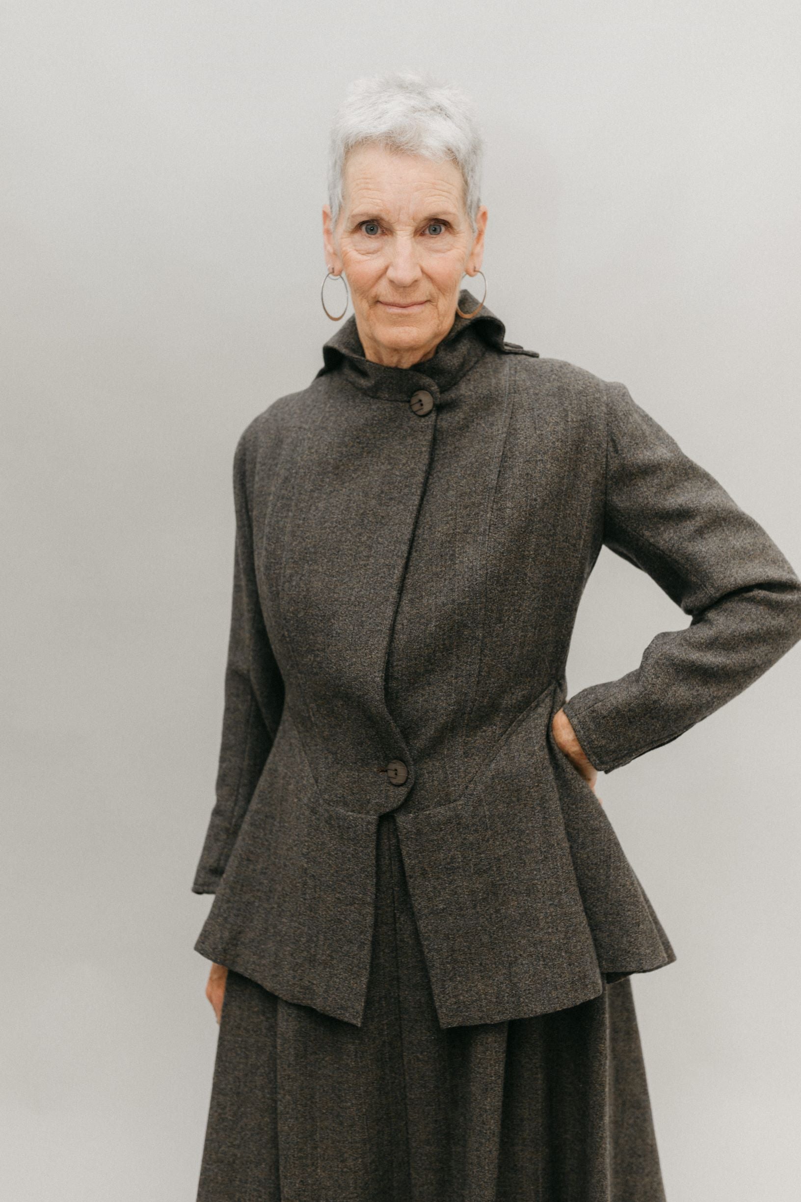 Older white woman with gray hair with her left hand on her hip, standing in front of a white studio backdrop wearing the traveling suit buttoned 