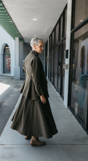 Older white woman with gray hair, mid twirl standing outside glass doors, shoulder and back facing front, the jumper is flared at the bottom.