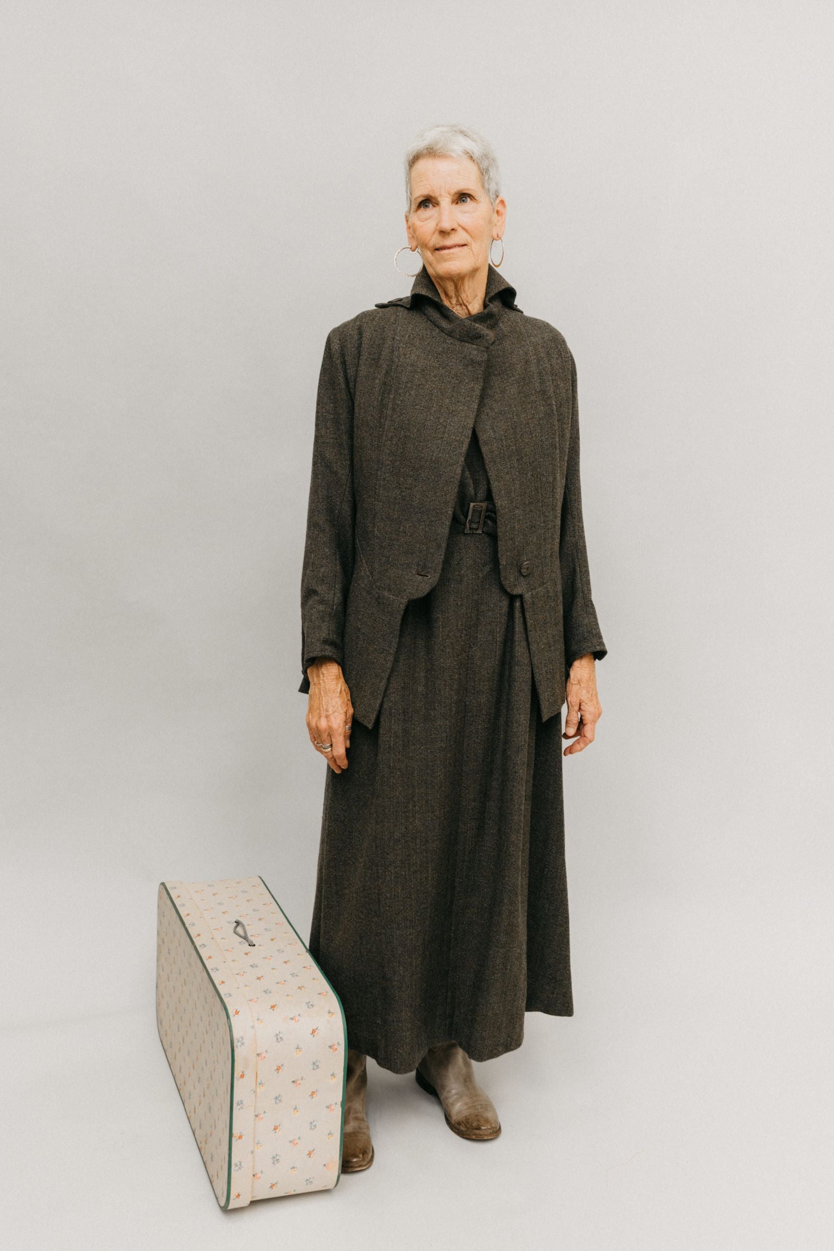 Older white woman, with gray hair wearing the 508 traveling suit, she is standing next to a white suitcase in front of a studio white backdrop